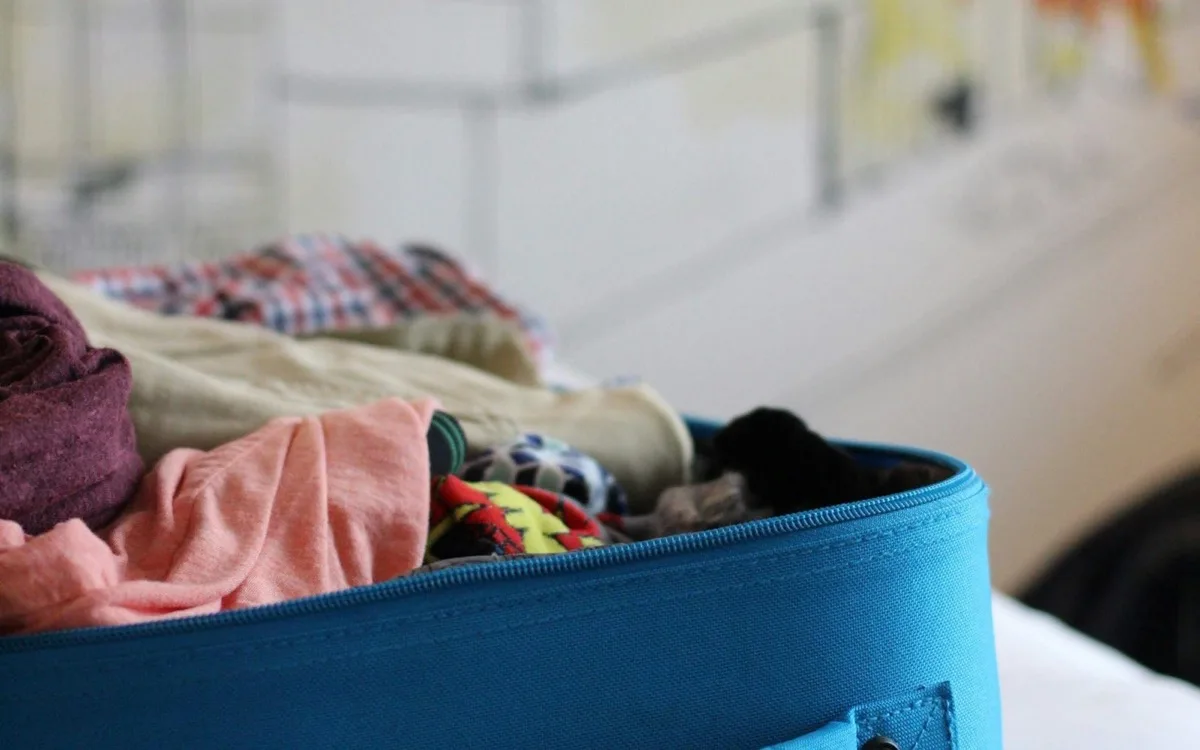 An open carry-on suitcase overstuffed with unfolded clothes on a bed