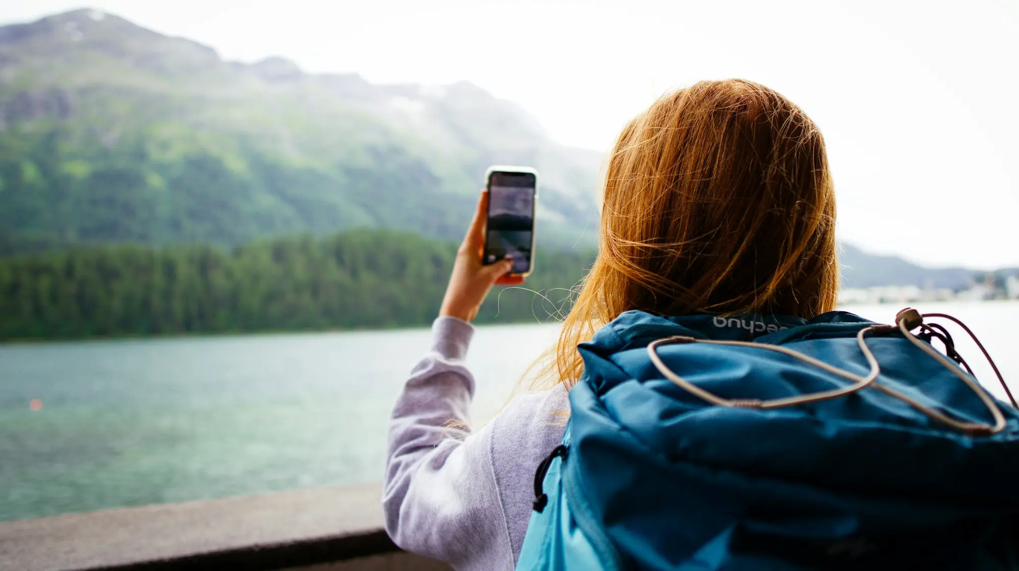 A hiker with a backpack holding a smartphone at a Swiss alpine lake with mountains behind