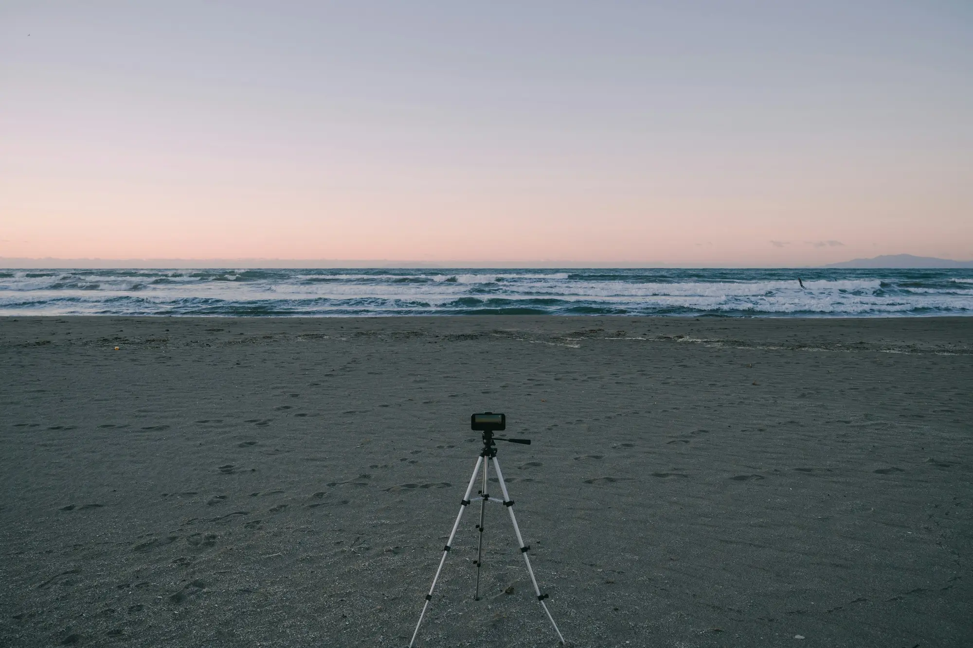 Camera tripod set up on a sandy beach at dusk facing the ocean waves